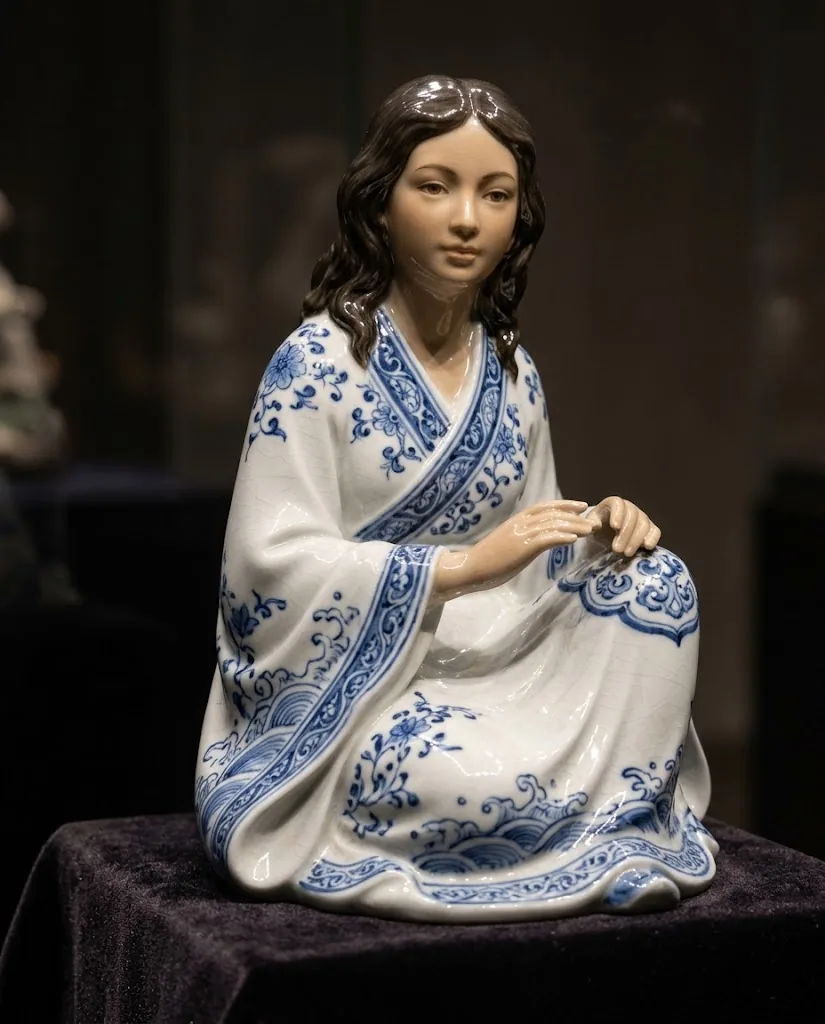A glossy white porcelain figurine of a seated woman in a robe painted with blue flowers and waves, on a dark velvet plinth.